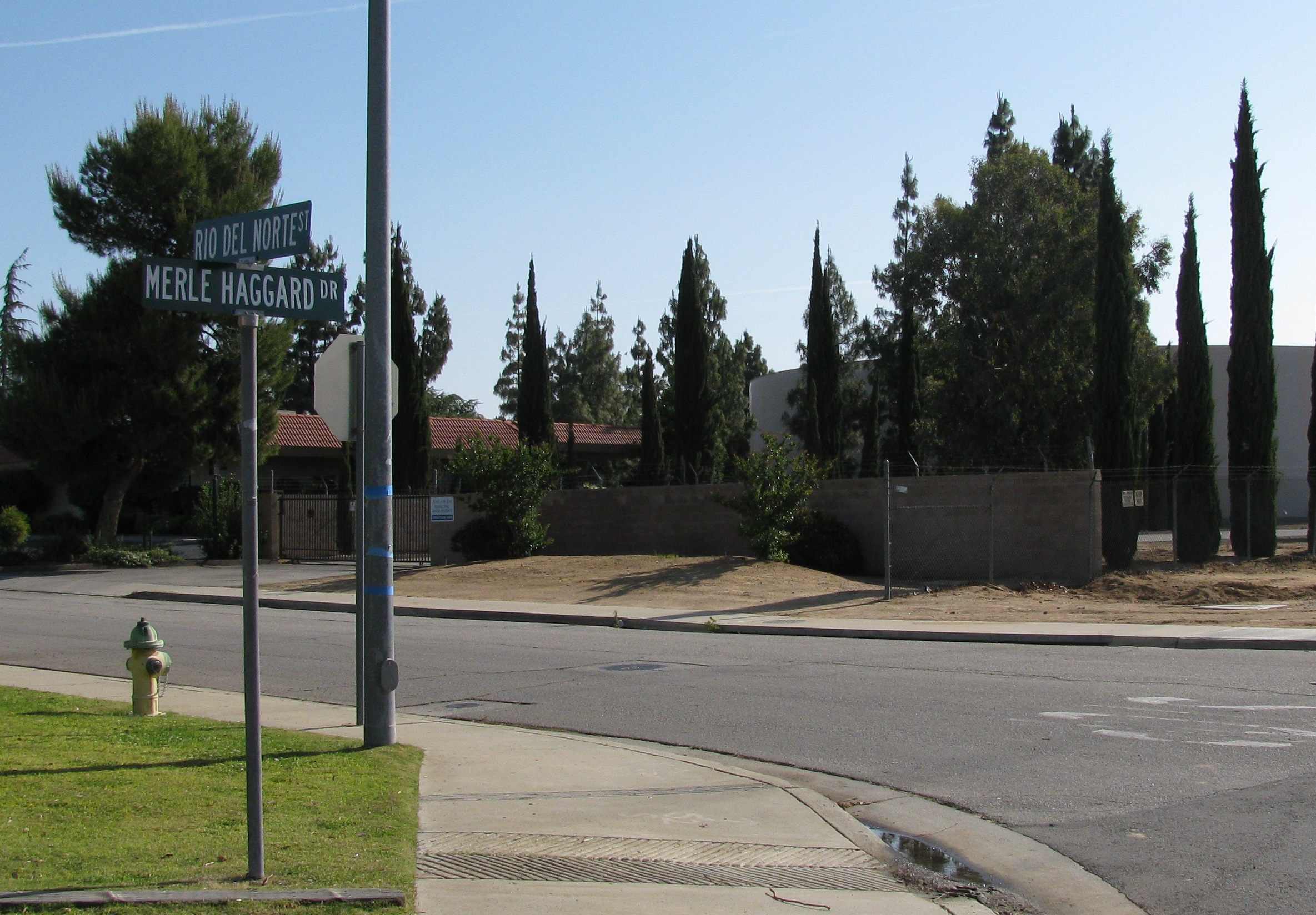 Street signs for Merle Haggard Drive and Rio Del Norte Street in Oildale, with trees and buildings behind them.