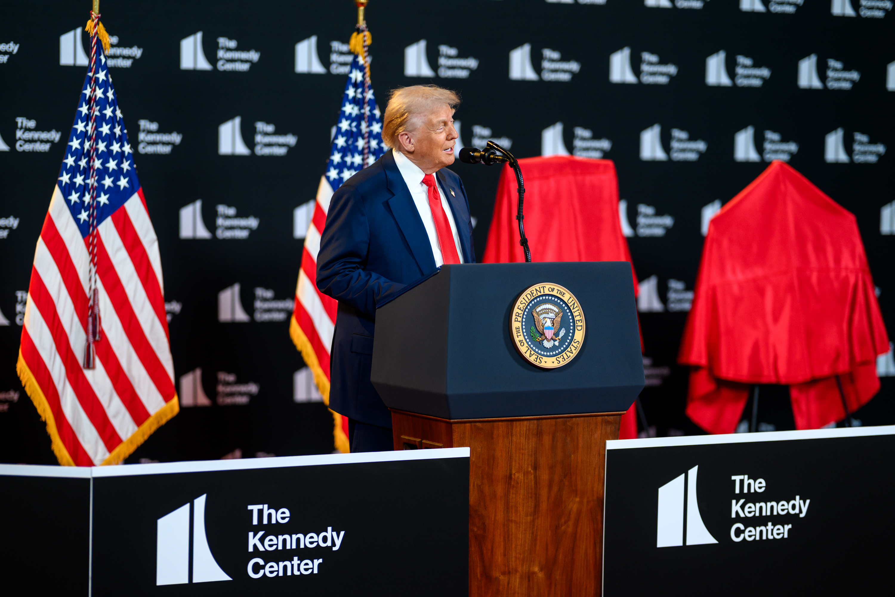 Donald Trump speaking at a presidential lectern, with U.S. flags behind him.