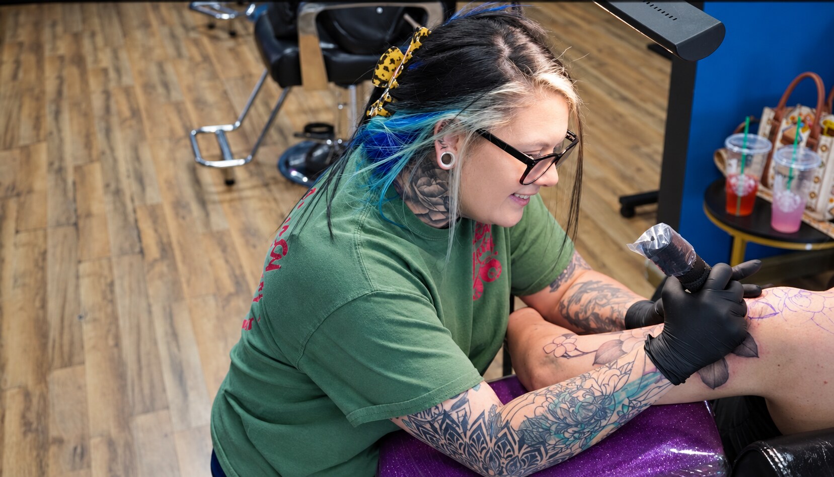 Tattoo artist Bre Bratton wearing black gloves and using a tattoo machine on a client's leg inside Voodoo Lily Tattoo in Bakersfield