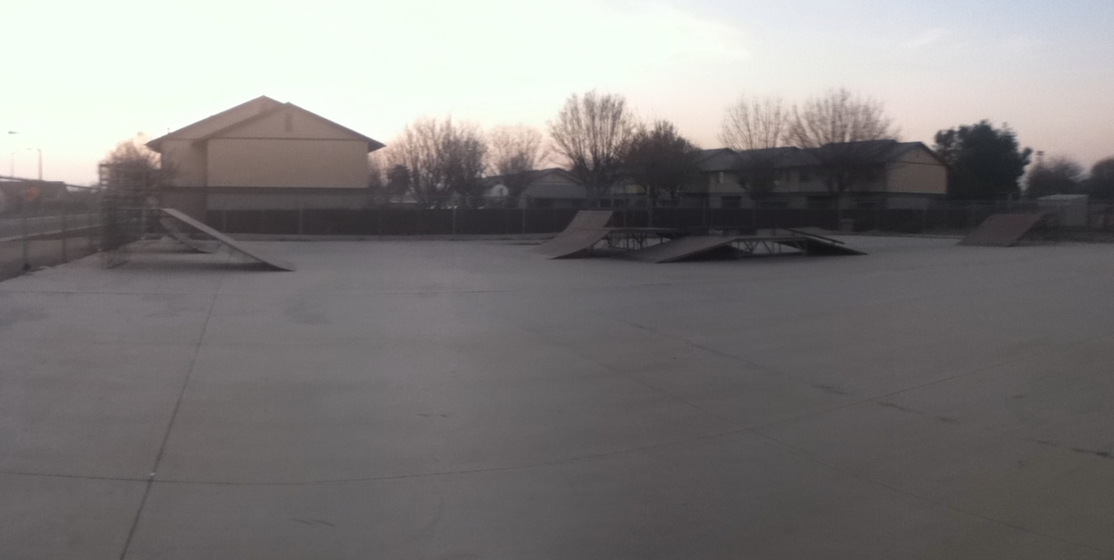 An empty concrete skate park in Shafter, with ramps and rails inside a fence and buildings and trees behind it.